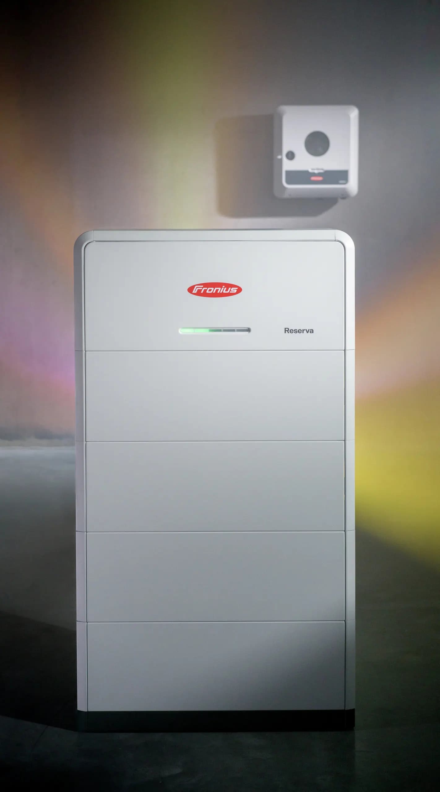 Fronius Reserva batteri på gulvet med en Fronius inverter på væggen over, mod en betonvæg i farvet lys