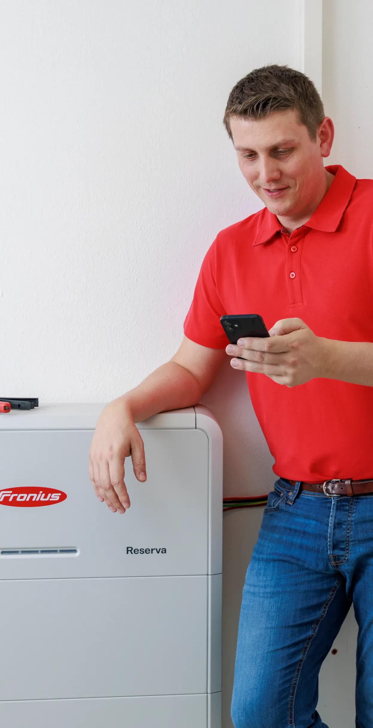 Installatør i rød Fronius-trøje kigger på sin telefon med armen hvilende på et Fronius Reserva batteri